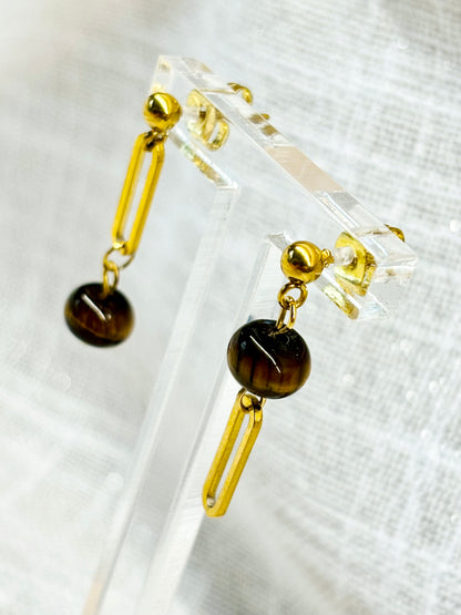 Boucles d'oreille NOËLA, avec pierre d'Oeil de Tigre, exposées sur un fond en lin.