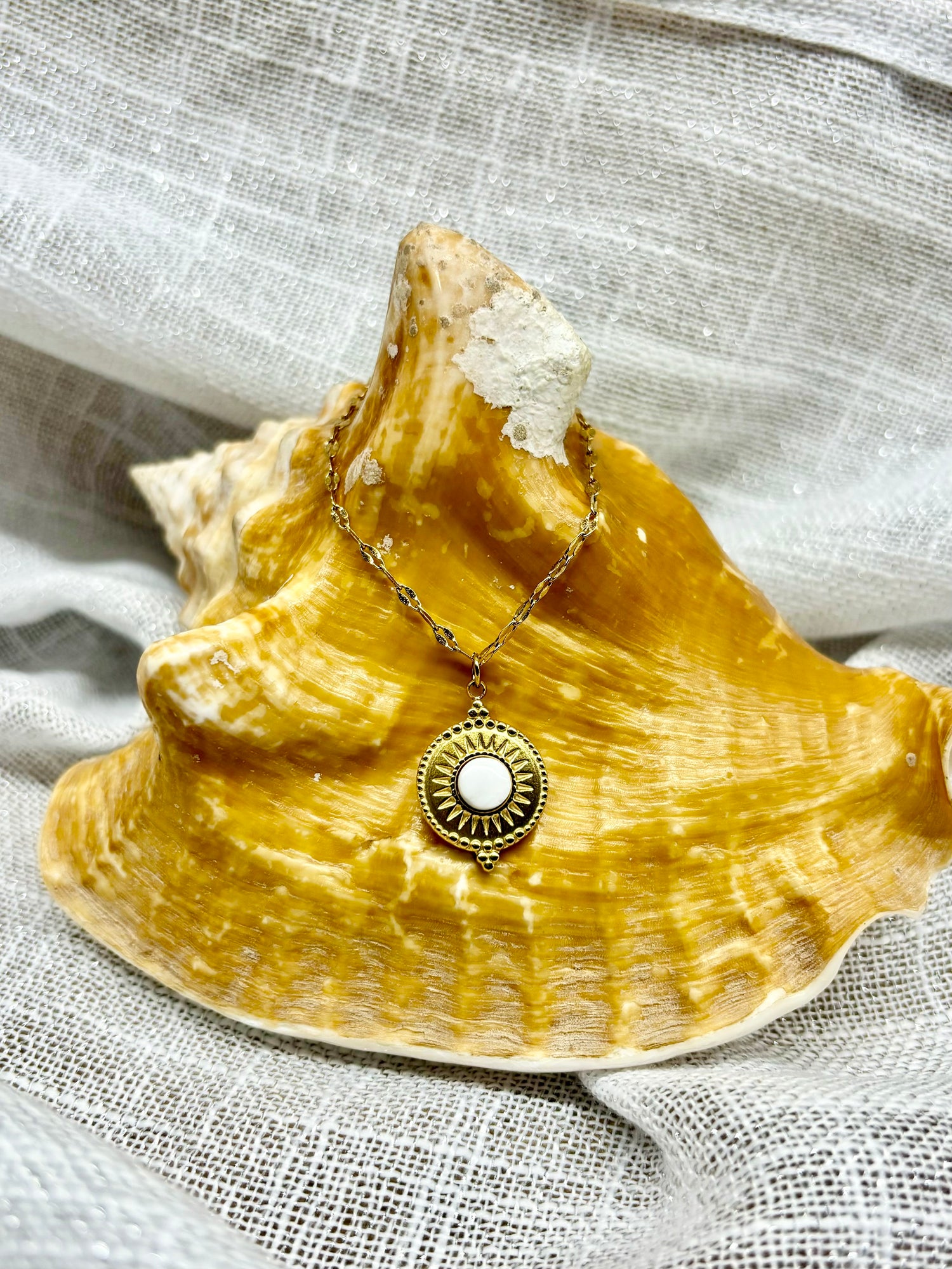 Collier ZÉLIE en acier inoxydable doré et pierre de Howlite, exposé sur un coquillage et un fond en lin. 