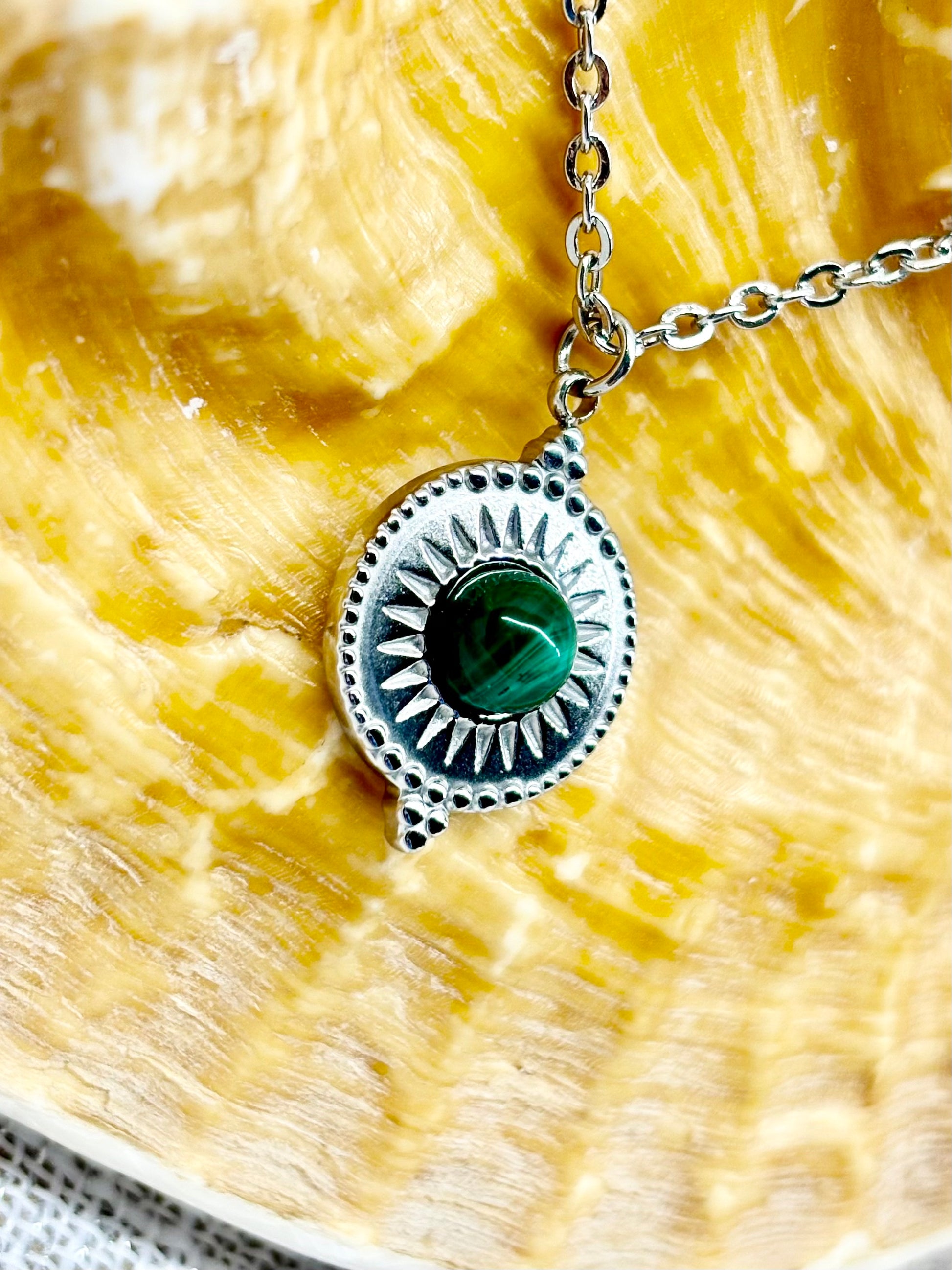 Gros plan du collier ZÉLIE en acier inoxydable argent et pierre de Malachite, exposé sur un coquillage et un fond en lin