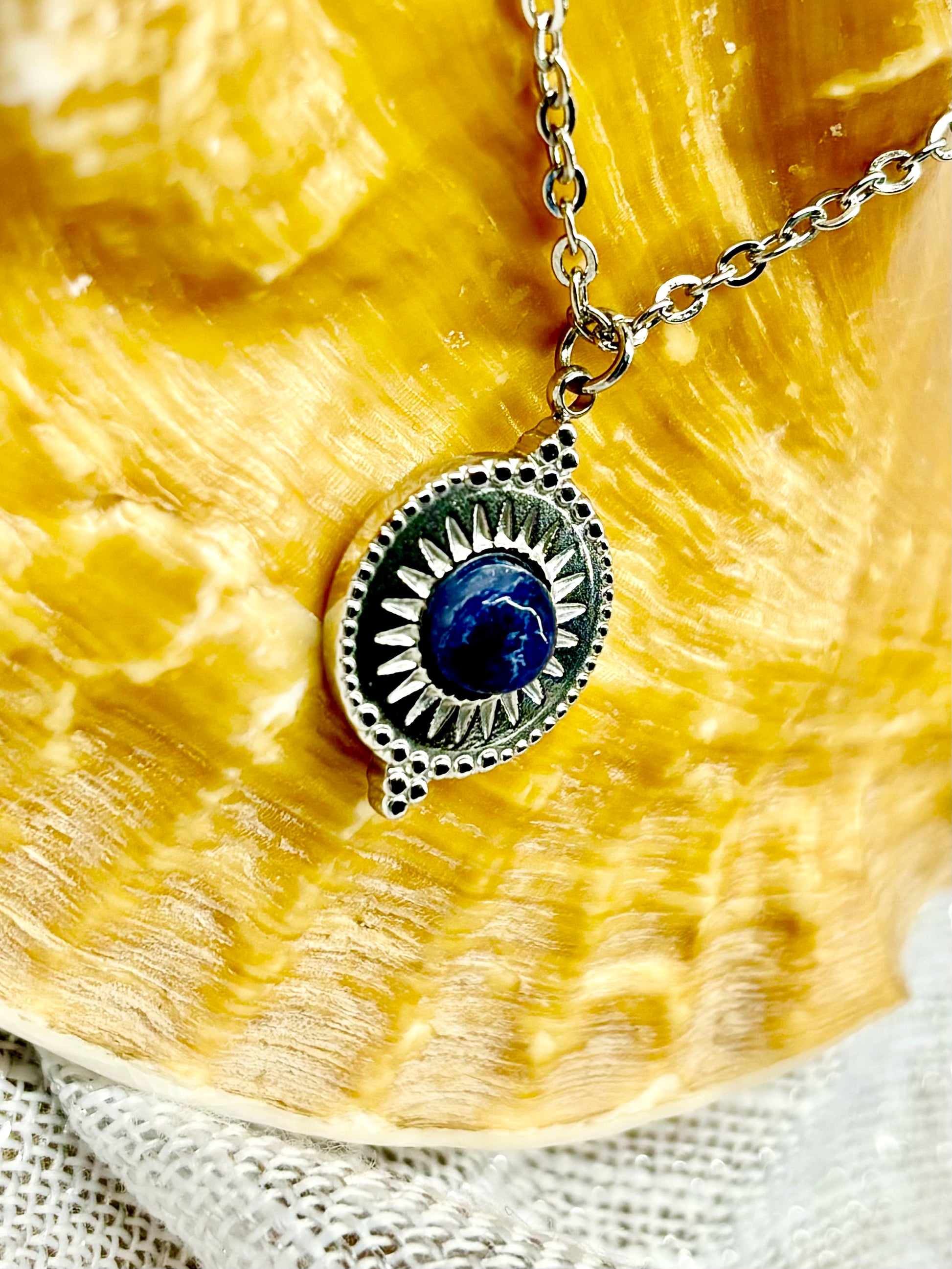 Gros plan du collier ZÉLIE en acier inoxydable argent avec une pierre de Sodalite, exposé sur un coquillage et un fond en lin.