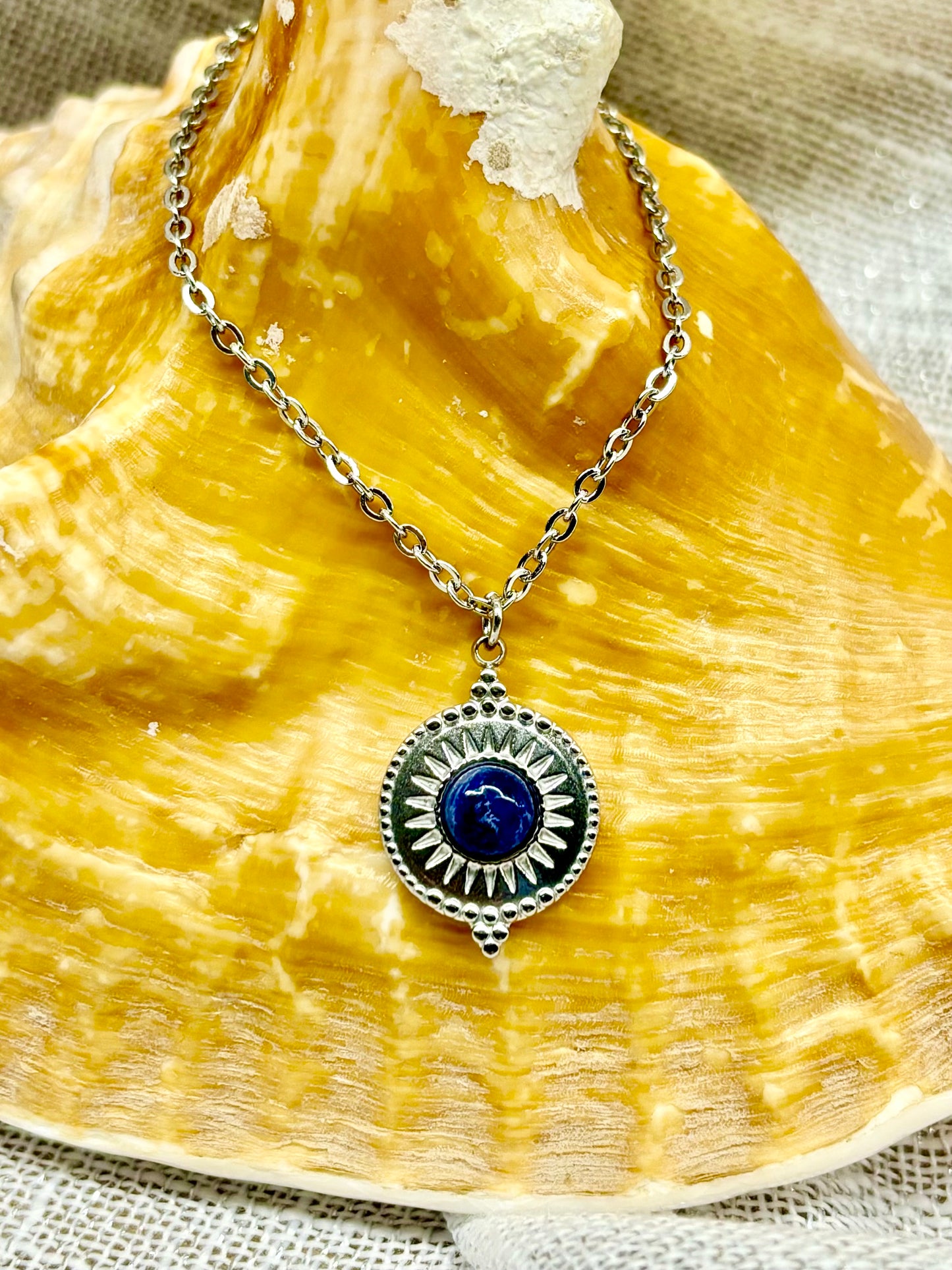 Collier ZÉLIE en acier inoxydable argent avec une pierre de Sodalite, exposé sur un coquillage et un fond en lin.