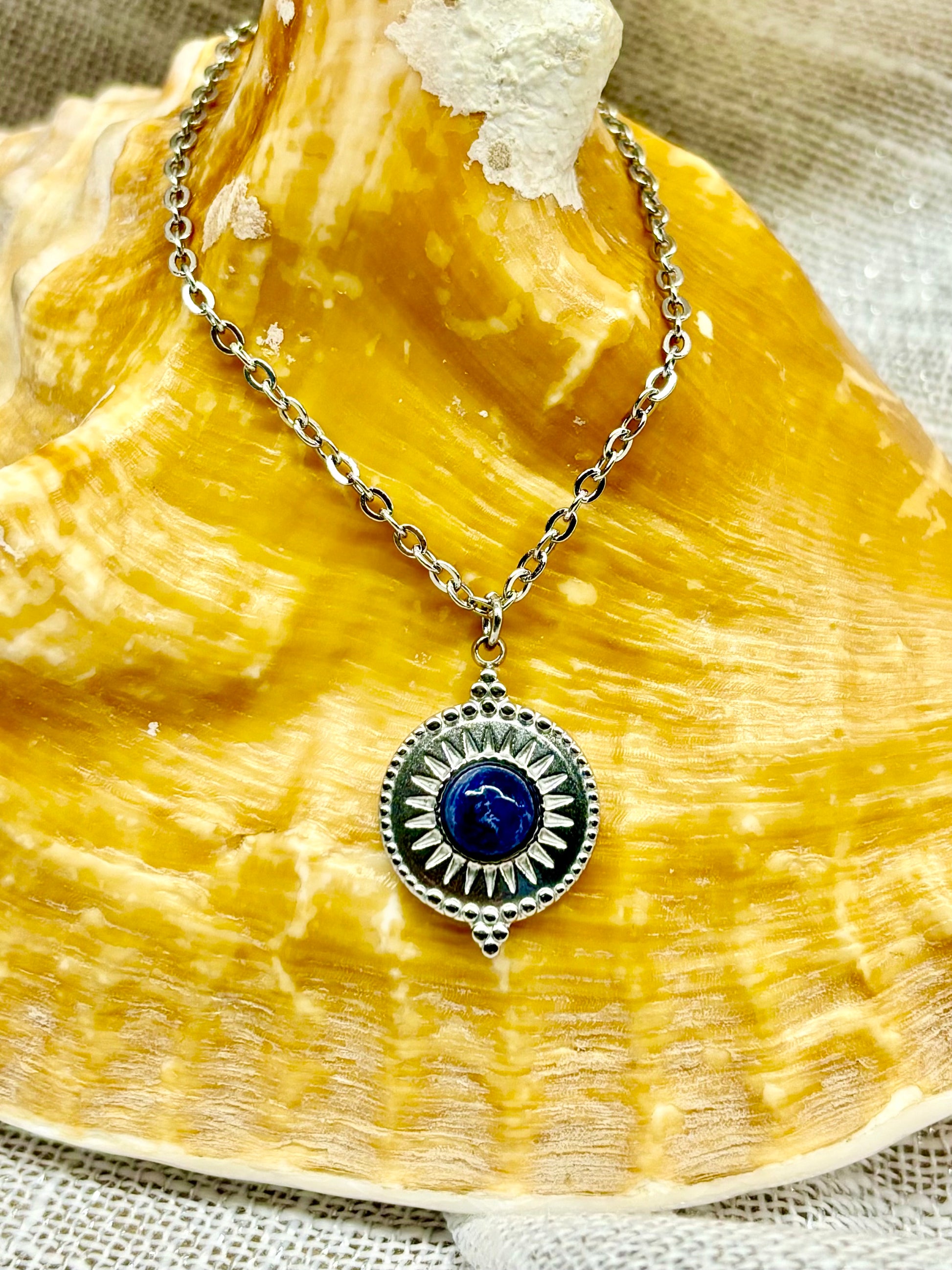 Collier ZÉLIE en acier inoxydable argent avec une pierre de Sodalite, exposé sur un coquillage et un fond en lin.