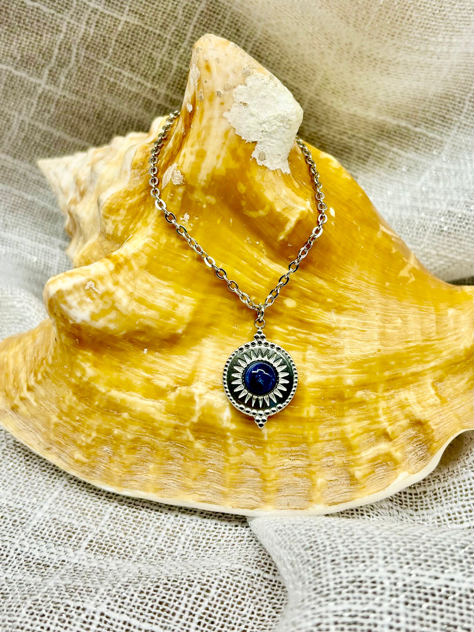 Collier ZÉLIE en acier inoxydable argent avec une pierre de Sodalite, exposé sur un coquillage et un fond en lin.
