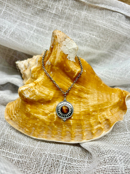 Collier ZÉLIE en acier inoxydable argent et pierre d'oeil de tigre, exposé sur un coquillage et un fond en lin