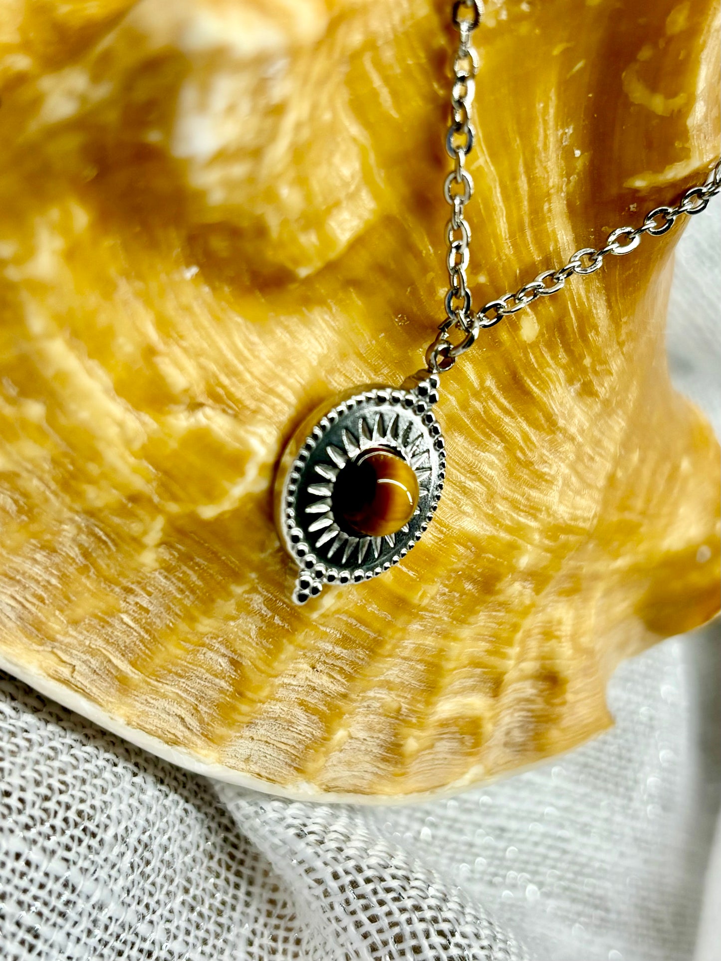 Gros plan du Collier ZÉLIE en acier inoxydable argent et pierre d'oeil de tigre, exposé sur un coquillage avec un fond en lin