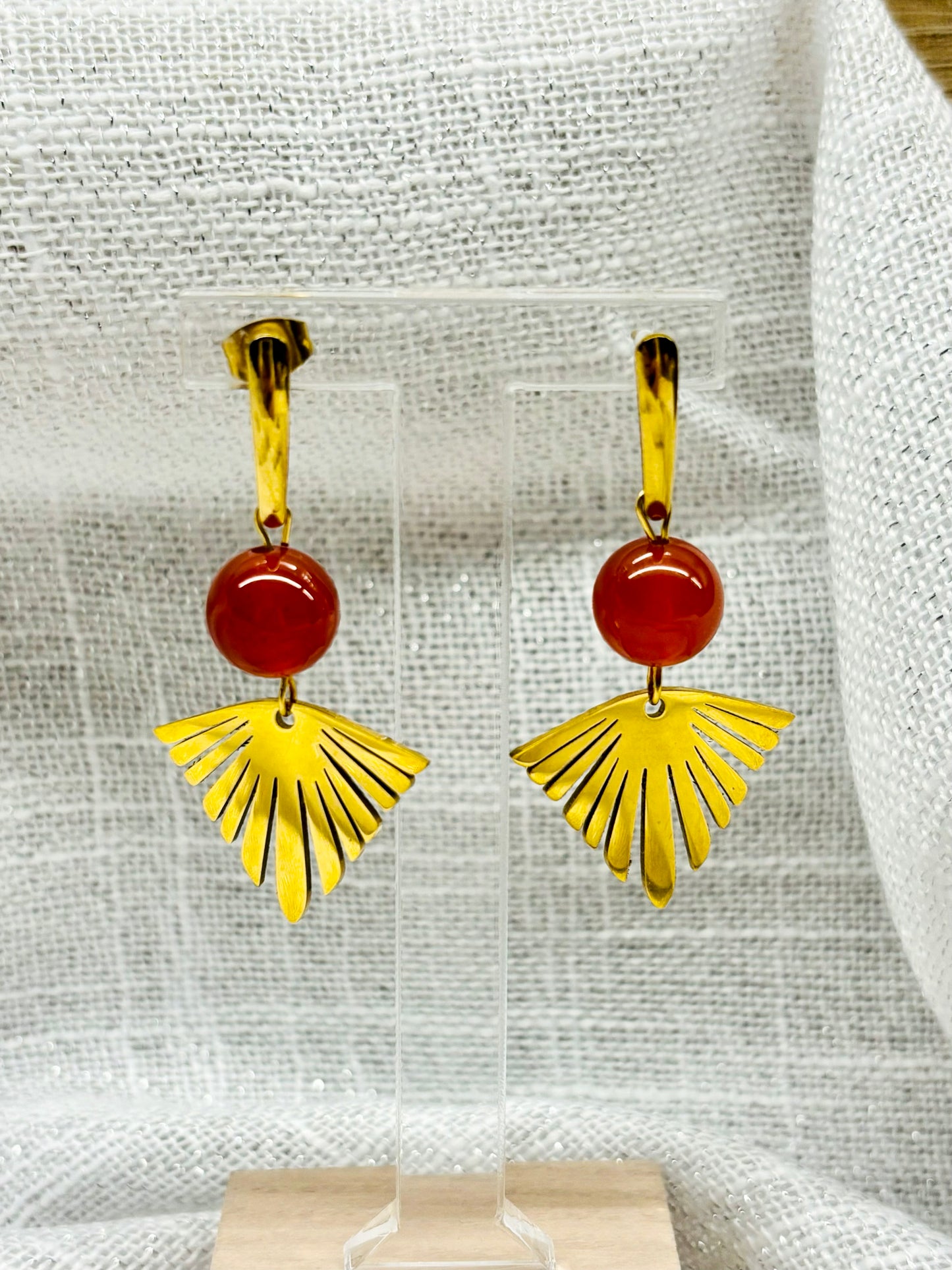 Paire de boucles d'oreille ZOA, en acier inoxydable doré et pierre de Cornaline, exposée sur un portant.