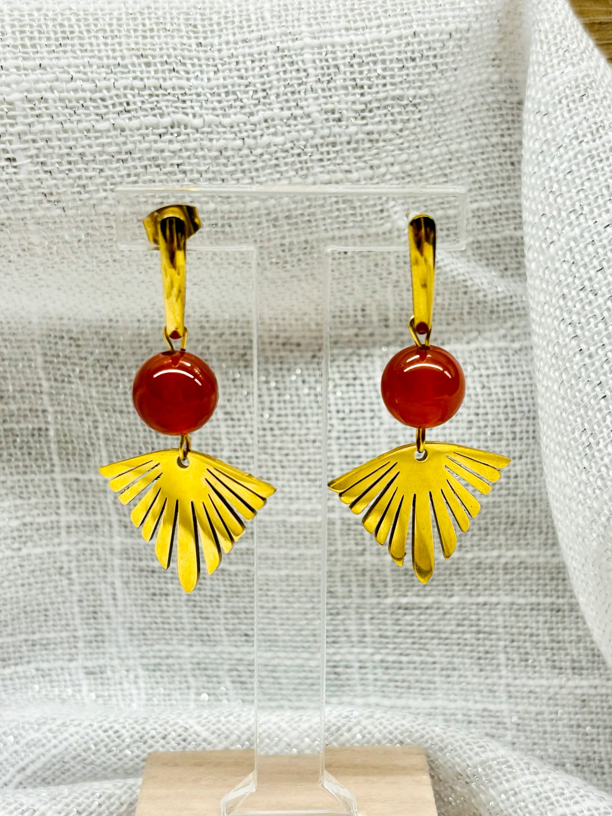 Paire de boucles d'oreille ZOA, en acier inoxydable doré et pierre de Cornaline, exposée sur un portant.